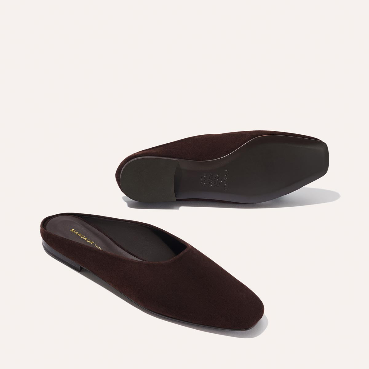 The Clara - Espresso Suede by Margaux: dark brown suede slip-ons with a slipper-like silhouette, squared toes, flat soles, and a minimalist design. One shoe is upright; the other lies on its side to show the sole.