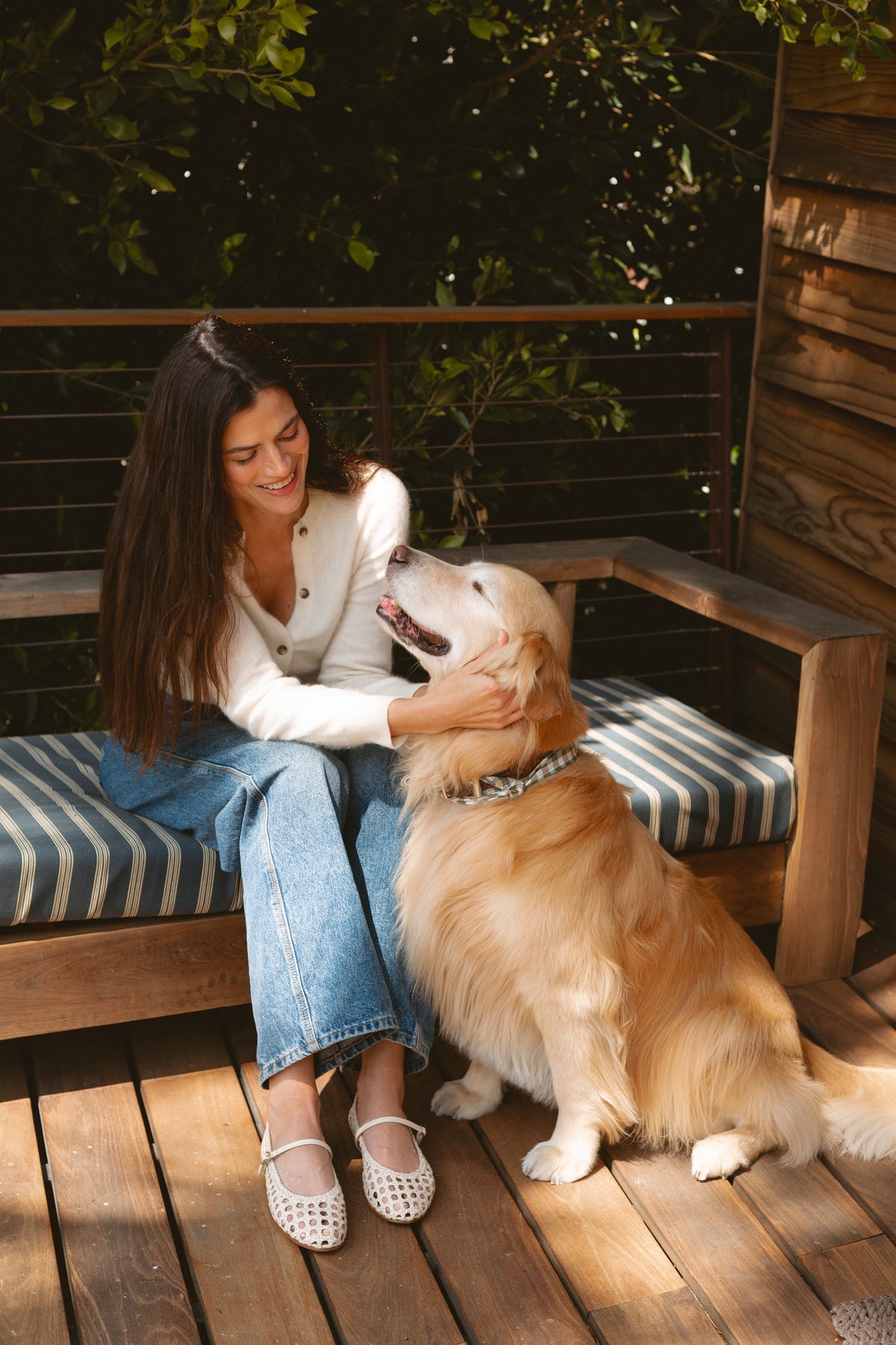 A woman with long brown hair, wearing a white sweater and jeans, sits on a striped outdoor bench, smiling and petting a happy golden retriever beside her on a wooden deck. Lush greenery is visible in the background.