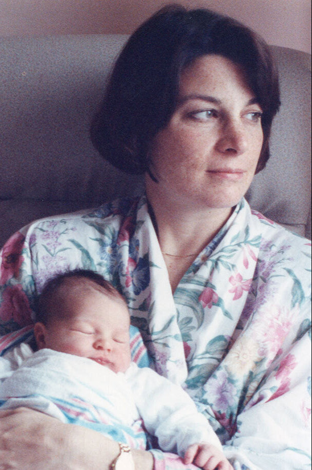A woman with short brown hair wearing a floral robe sits on a chair, looking softly to the side, while cradling a sleeping baby wrapped in a blanket in her arms.