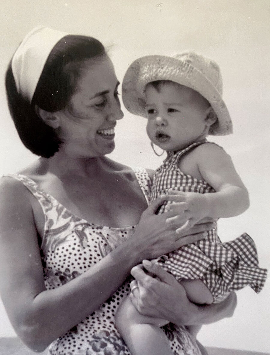 A woman in a sleeveless dress and headband holds a baby in a checkered romper and sunhat. Both are smiling and looking at each other. The photo is in black and white.