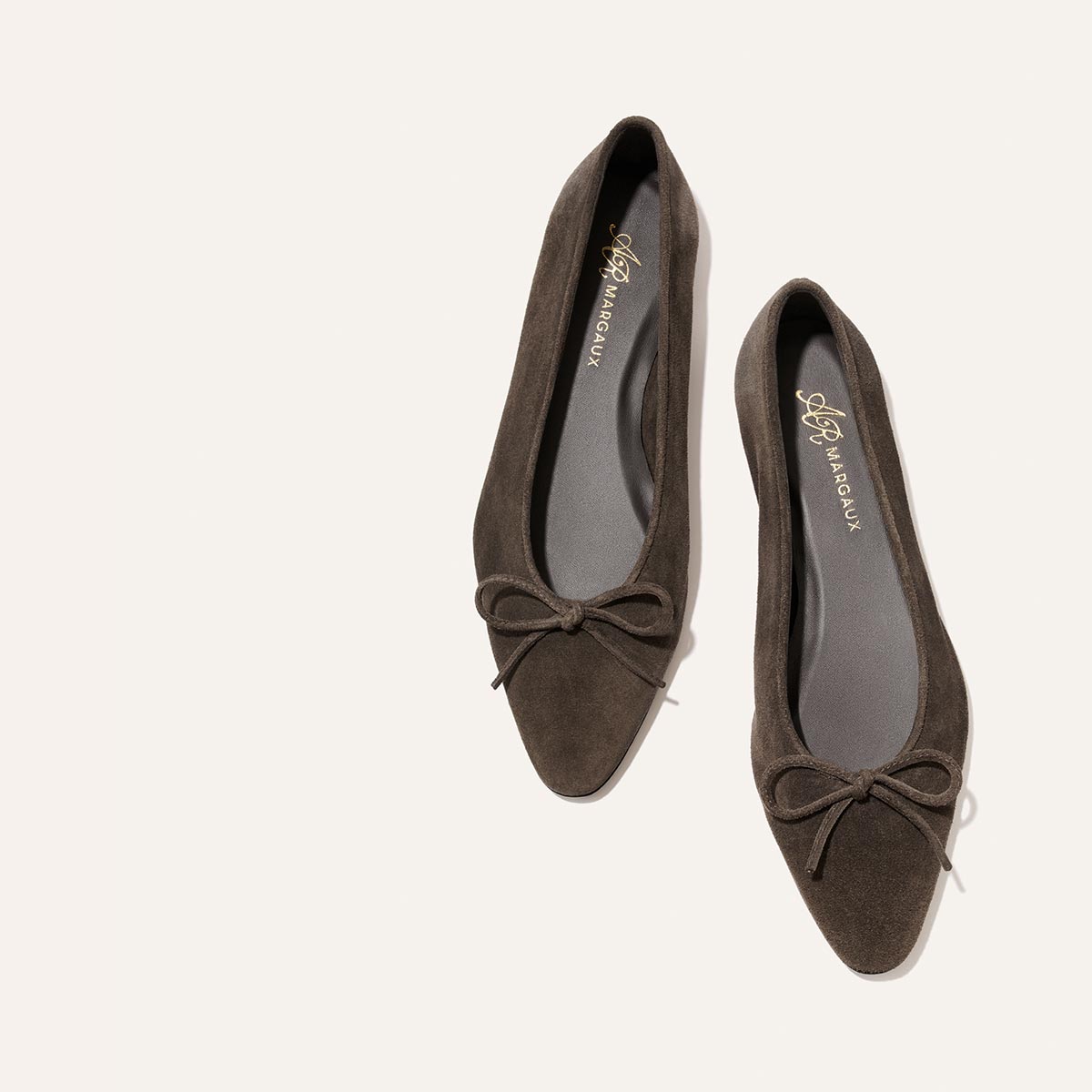 A pair of Alison Roman x Margaux: The Salamanca flats in English Brown suede by Margaux, featuring pointed toes and bows, are shown from above on a light background.