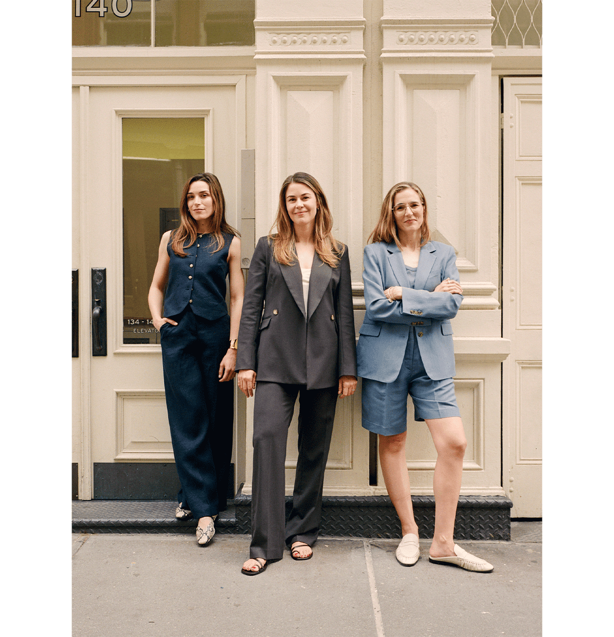 Three women stand in front of a tan building entrance, each wearing stylish business attire: a dark vest and pants, a dark double-breasted suit, and a blue blazer with matching shorts and loafers. They look confidently at the camera.