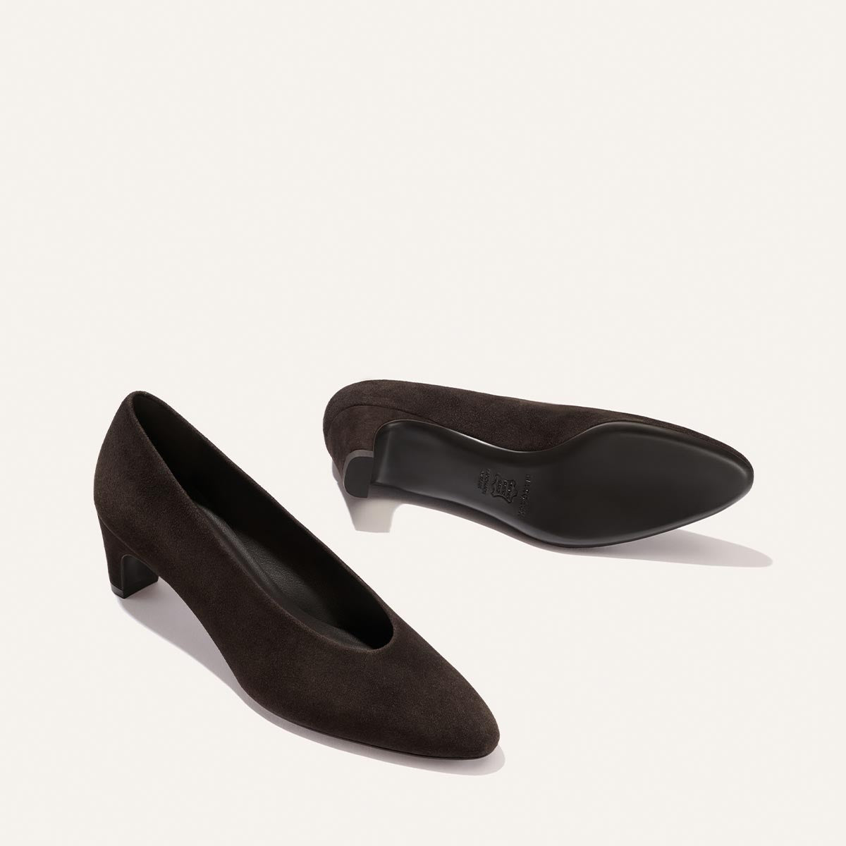 A pair of The Augusta Glove Pump in Molasses Suede by Margaux, one shoe upright and the other on its side to display the sole, set against a plain off-white background.