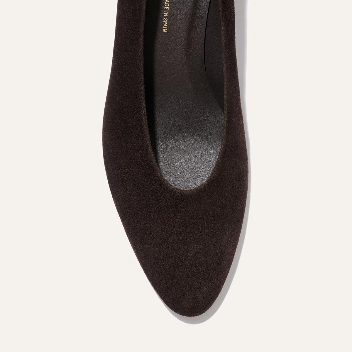 A close-up top view of Margaux’s The Augusta Glove Pump in Molasses Suede, a dark brown pointed-toe flat, on a light background.