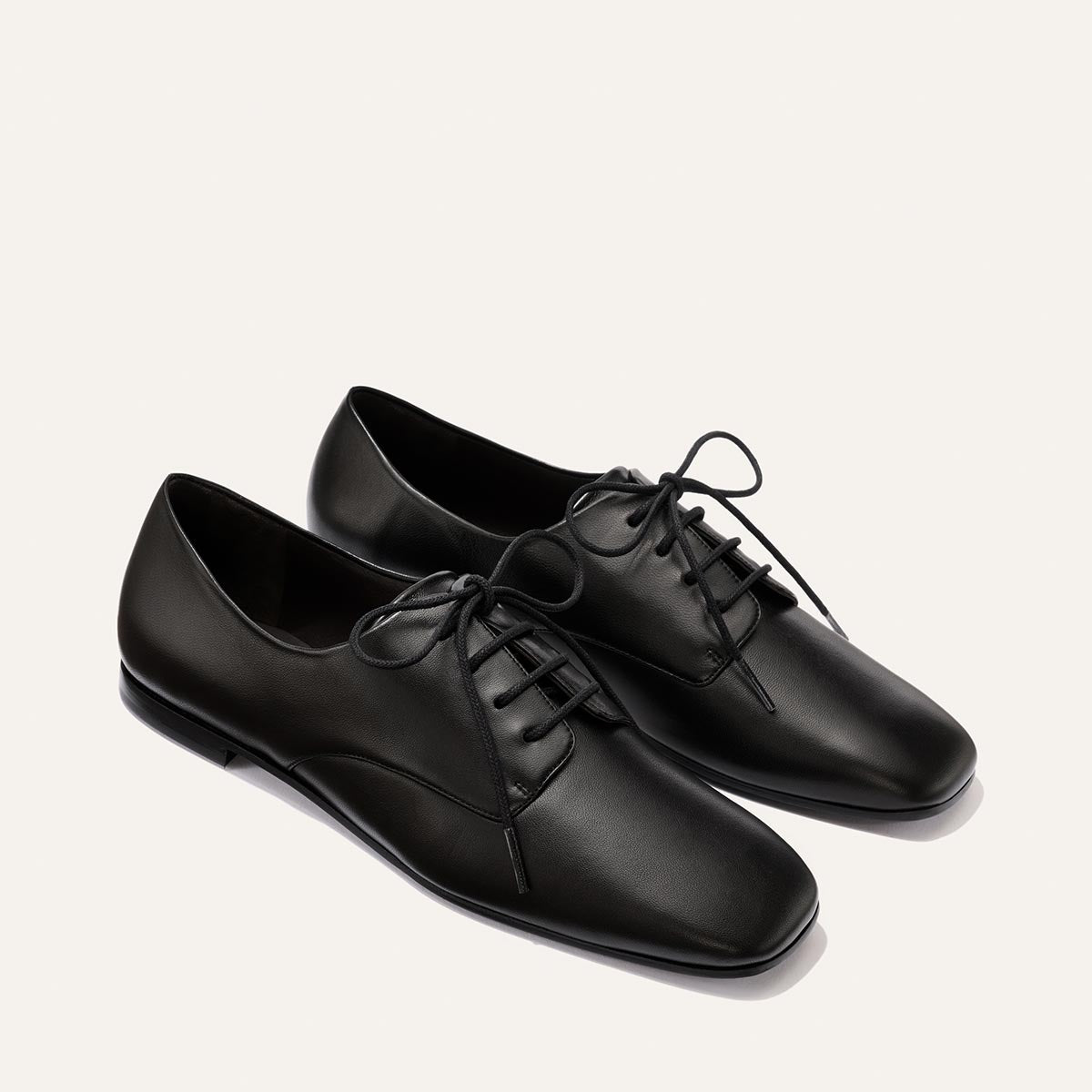 The Billie Derby - Black Nappa by Margaux: a pair of black leather lace-up dress shoes featuring a sleek, minimalist design and slightly squared toes, displayed on a plain, light background.