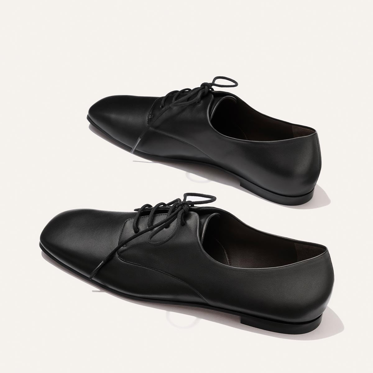 A pair of Margaux’s The Billie Derby shoes in Black Nappa leather is displayed on a light background, with one shoe positioned slightly above the other.