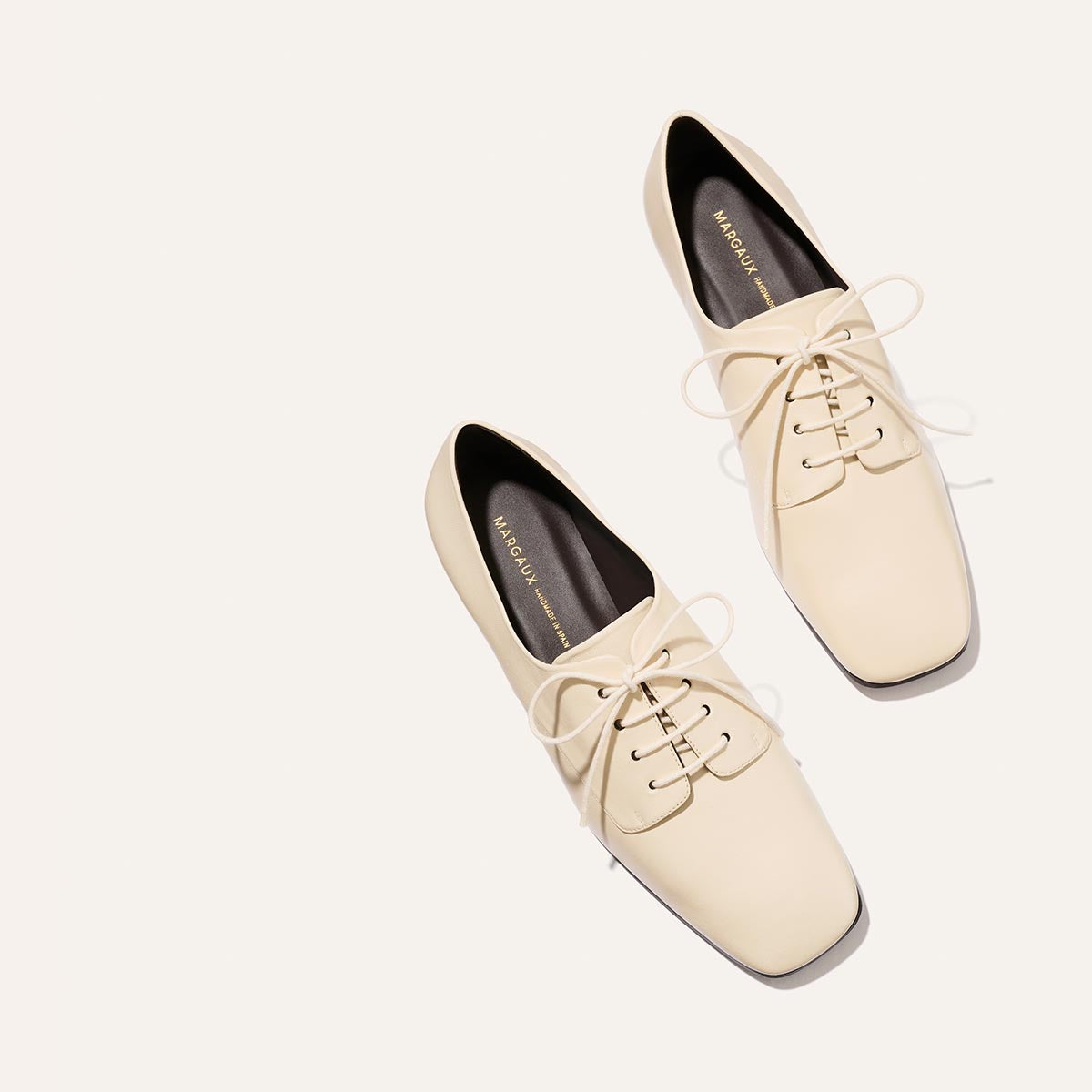 A pair of The Billie Derby - Vanilla Nappa oxford shoes by Margaux, featuring a cream-colored lace-up design with black interiors, is arranged diagonally on a plain light background.