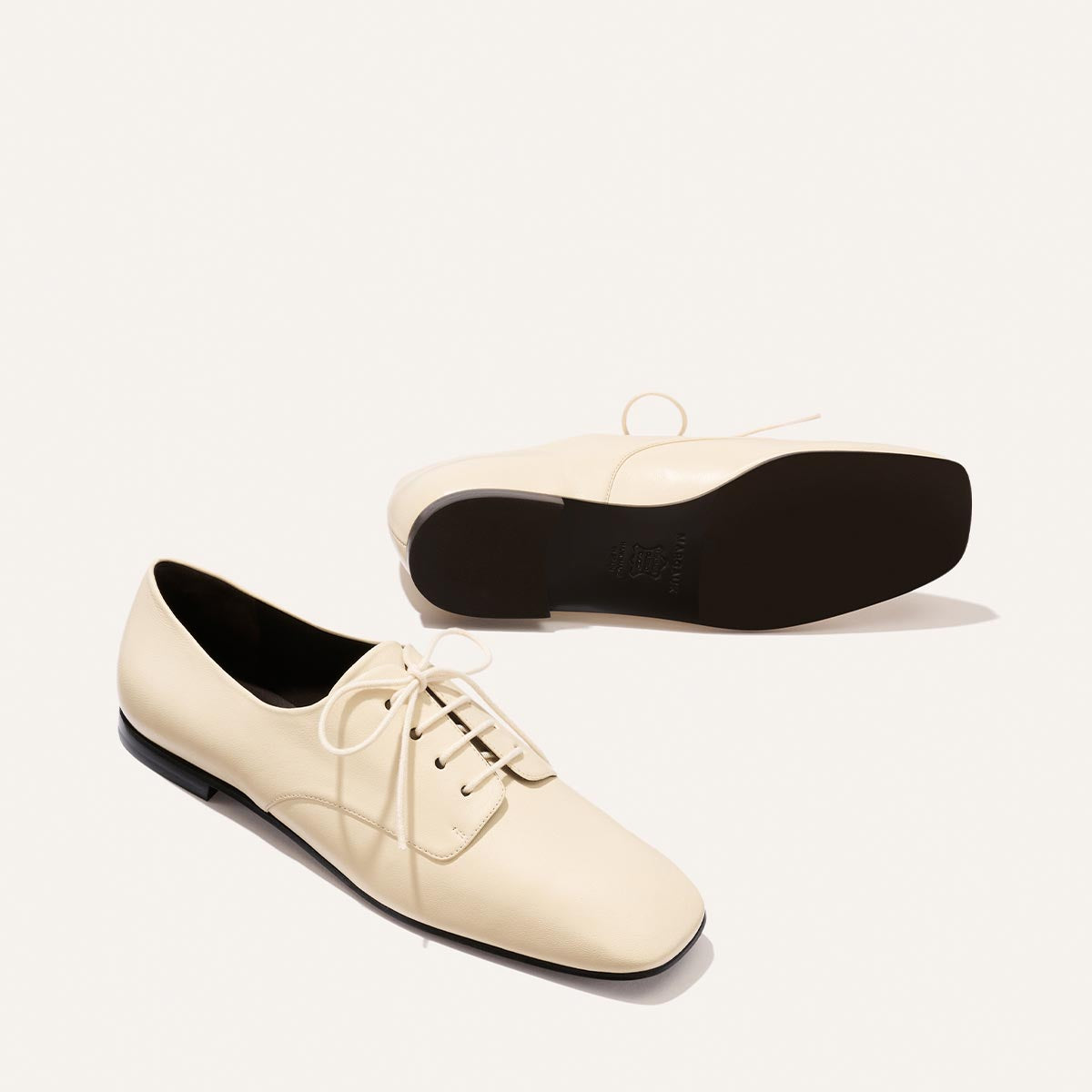 A pair of Margaux's The Billie Derby - Vanilla Nappa shoes: cream-colored lace-up leather with black soles, one shoe lying on its side to display the sole, the other standing upright.