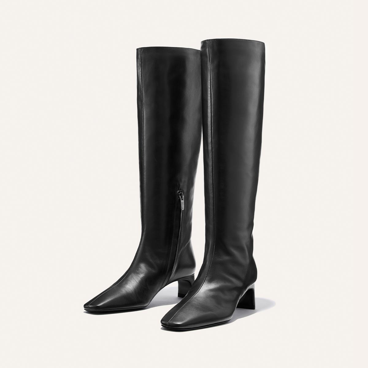 A pair of Margaux’s The Brera Knee-High Boot in Black Calf—sleek black leather boots featuring side zippers, pointed toes, and low block heels—are positioned upright on a plain light background.
