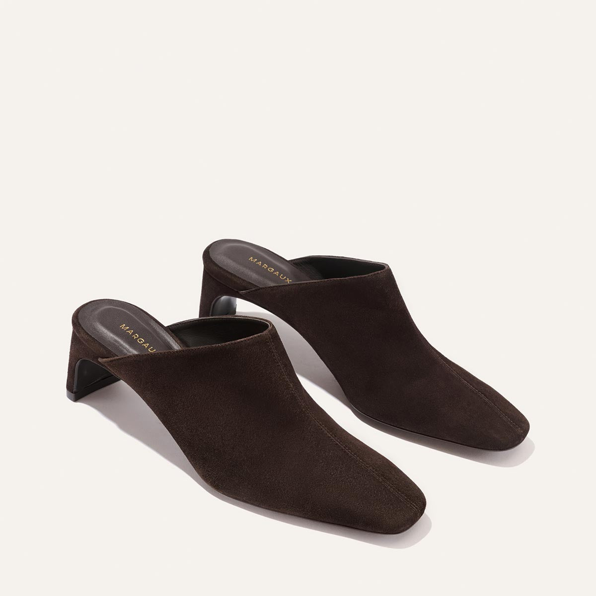 The Brera Mule - Molasses Suede by Margaux, a pair of dark brown suede mules with square toes, a high vamp, and mid-height block heels for an elegant silhouette, displayed on a light background.
