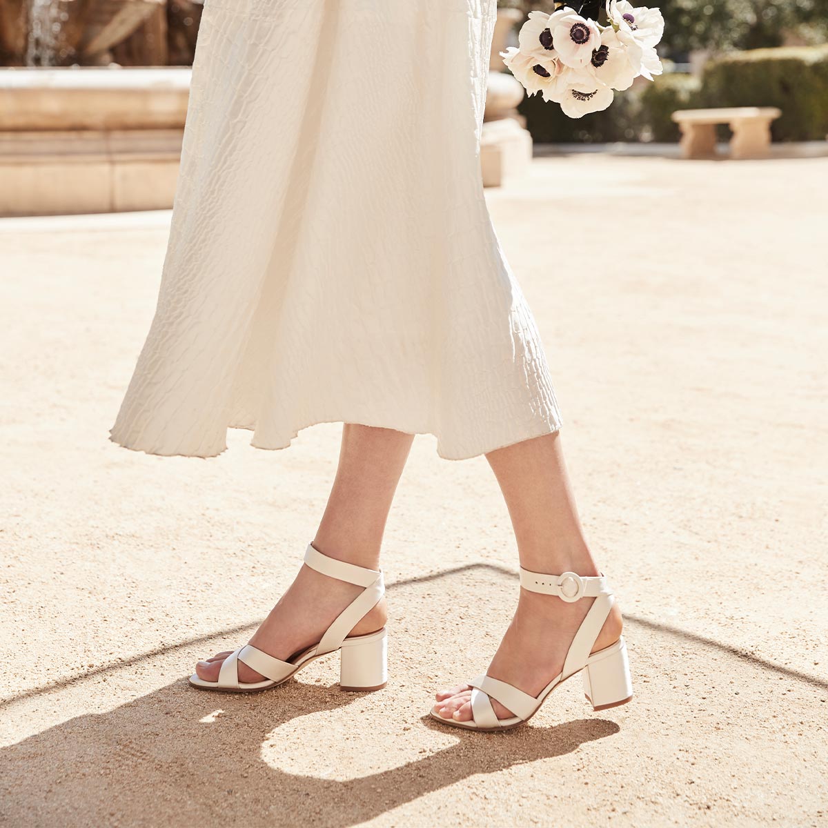 A woman in a white dress and Margaux’s bespoke bridal shoes strolls along a sandy path, holding a small bouquet—celebrate her special day with The Bridal Atelier E-Gift Card from Margaux.