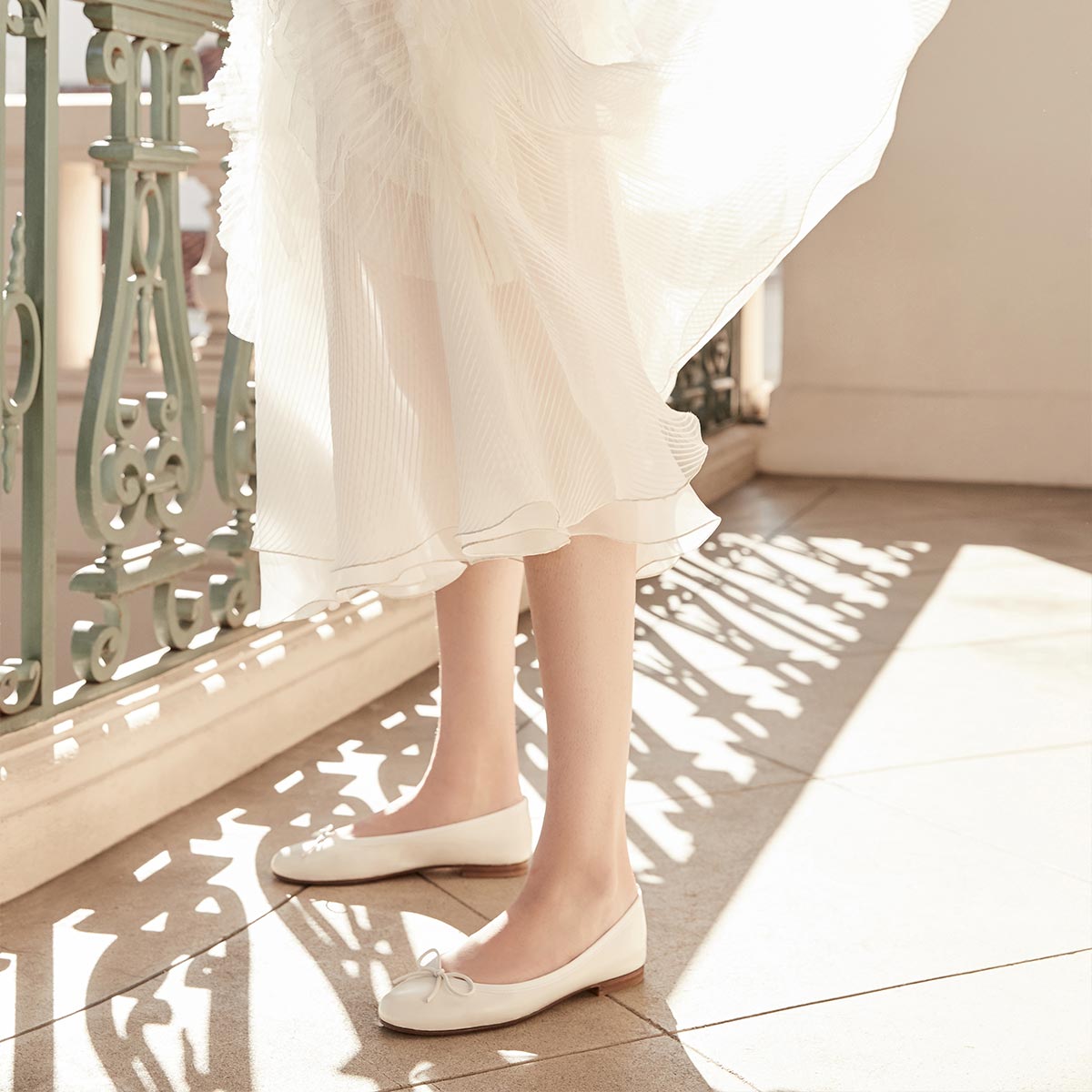 A person holds The Bridal Atelier E-Gift Card by Margaux on a sunlit balcony, their flowing white dress and bespoke bridal shoes casting soft shadows near an ornate green railing.