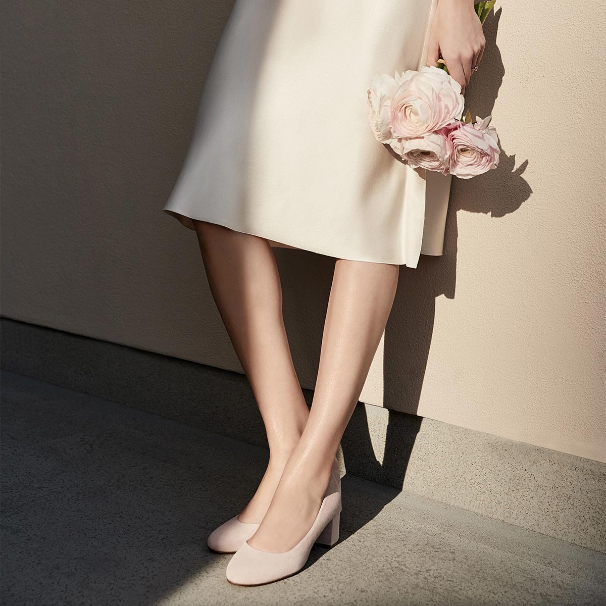 A woman in a light dress stands against a beige wall, holding pale pink flowers. Only her legs and dress show, paired with bespoke bridal shoes. Her look is completed with The Bridal Atelier E-Gift Card from Margaux.