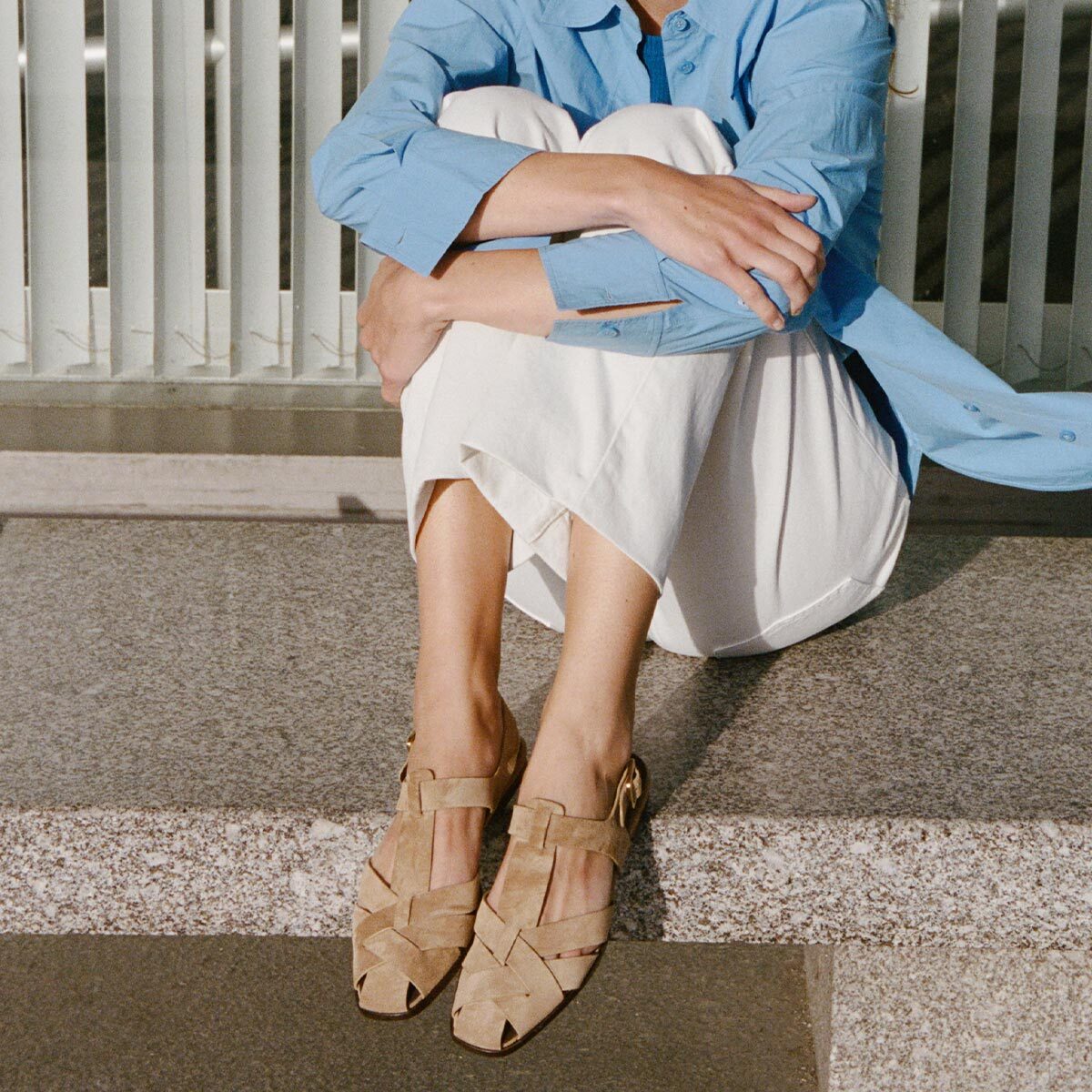 Person wearing a blue shirt, white skirt, and beige woven shoes sitting on steps.