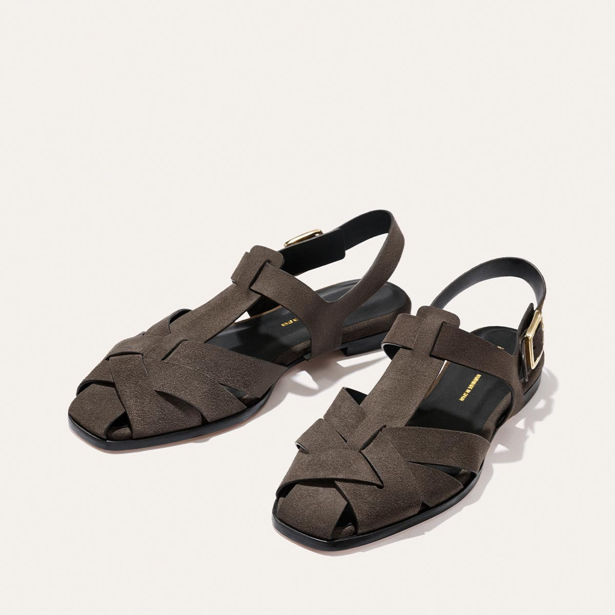 The Calypso Fisherman by Margaux in English Brown Suede features a modern interwoven pattern, slingback design, gold buckle closures, and flat soles, shown on a light background.