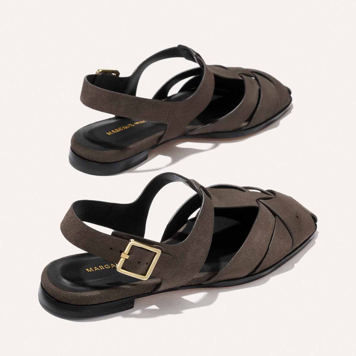 Two Calypso Fisherman sandals by Margaux in English Brown suede, featuring crossover interwoven straps, open toes, gold buckles, flat black soles, and the brand name in gold on the footbed. Displayed on a light background.