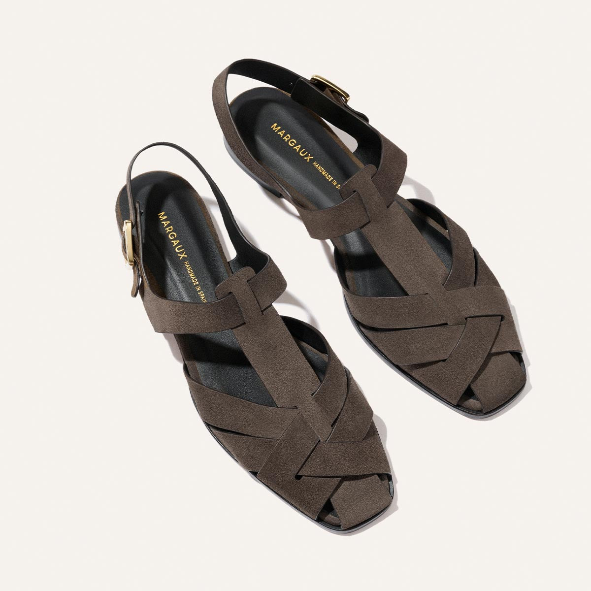 The Calypso Fisherman by Margaux in English Brown Suede features a modern interwoven design, open toes, slingback strap with gold buckle, and black insoles with gold branding—displayed on a light background.