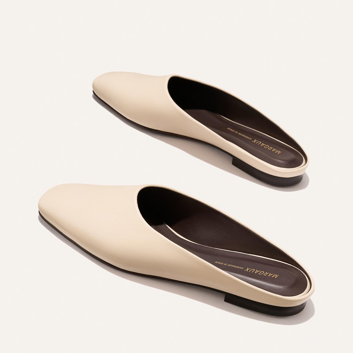The Clara - Bone Nappa by Margaux: beige slip-on leather mules with flat black soles, dark brown insoles, and gold branding, showcased on a light background.