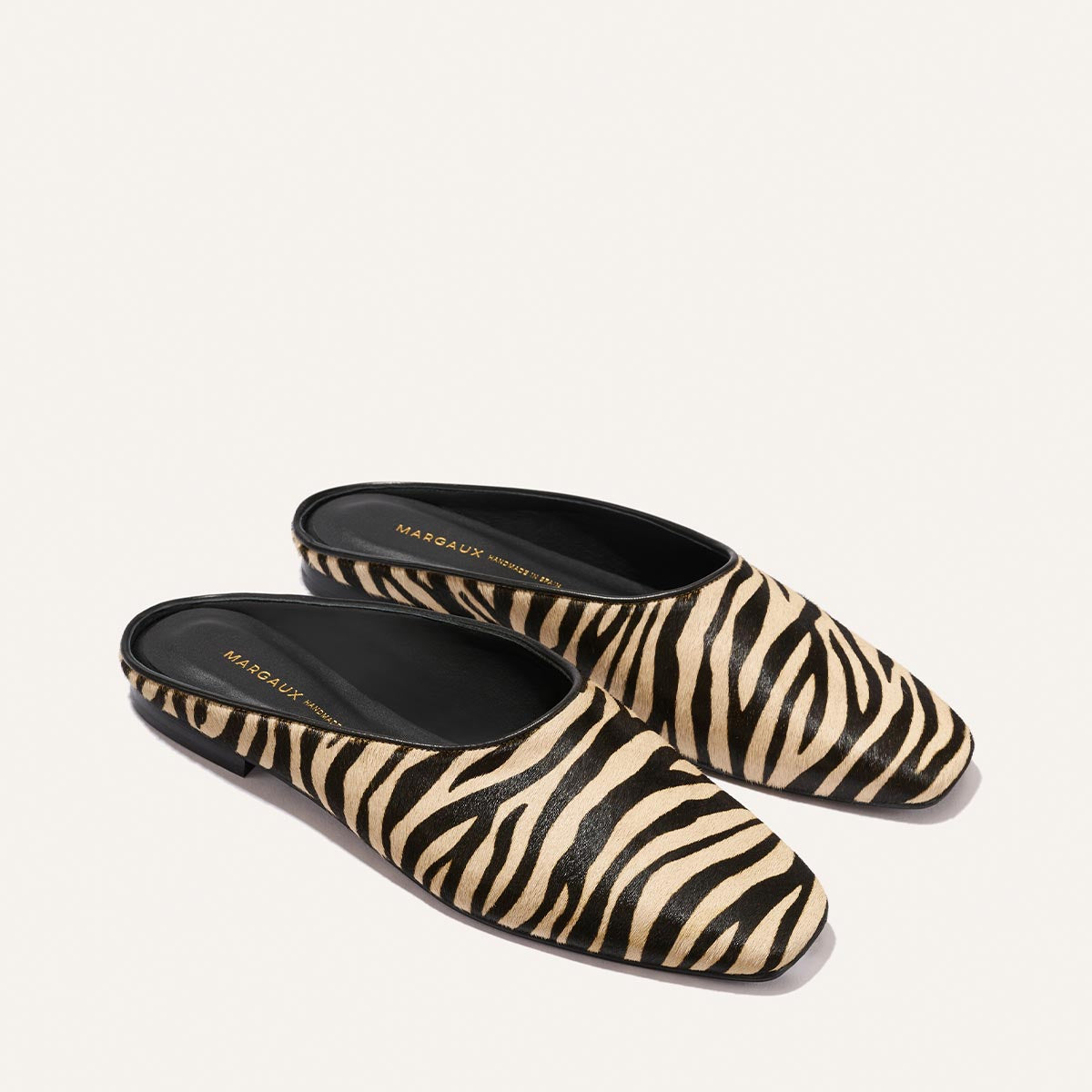 The Clara - Tan Zebra Haircalf by Margaux: slip-on mule shoes featuring a black and beige zebra print with black soles, shown on a light background.