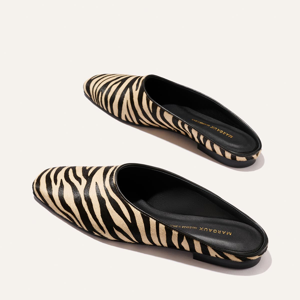 The Clara - Tan Zebra Haircalf by Margaux: stylish slip-on shoes featuring a black and beige zebra print, black insoles, and pointed toes, shown against a light background.