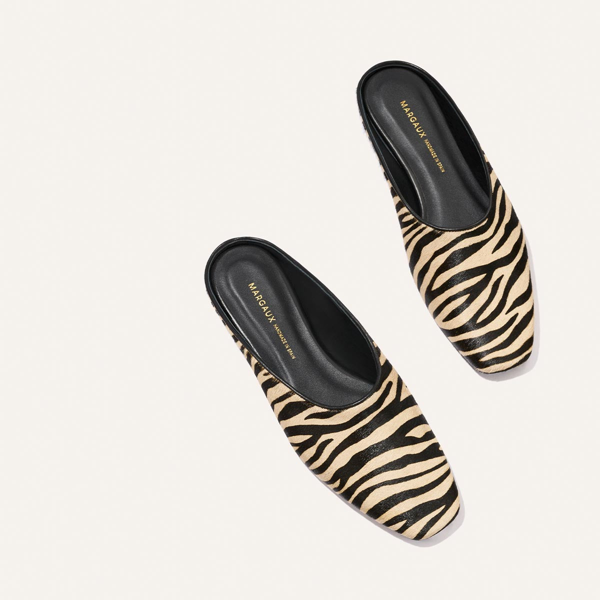 A pair of The Clara - Tan Zebra Haircalf slip-on mule shoes by Margaux, featuring a tan and black zebra stripe pattern with black insoles and Margaux branding in gold on the footbed, shown on a white background.