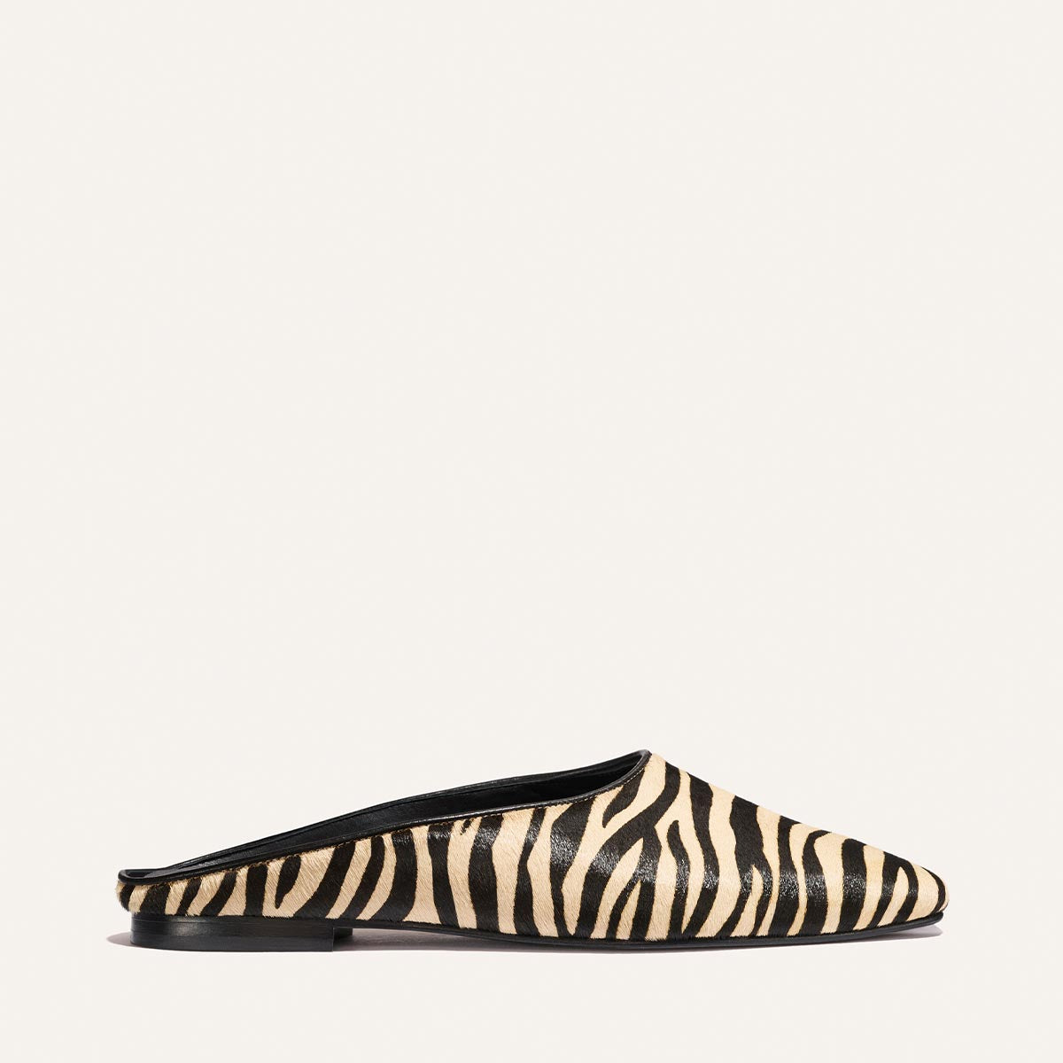 The Clara - Tan Zebra Haircalf by Margaux is a slip-on mule featuring a black and beige zebra print, flat sole, and black trim, shown against a plain light background.