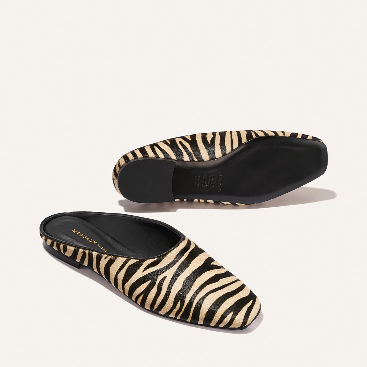 The Clara - Tan Zebra Haircalf by Margaux: slip-on mules with a tan and black zebra print, square toe, and flat black soles, shown on a plain white background.