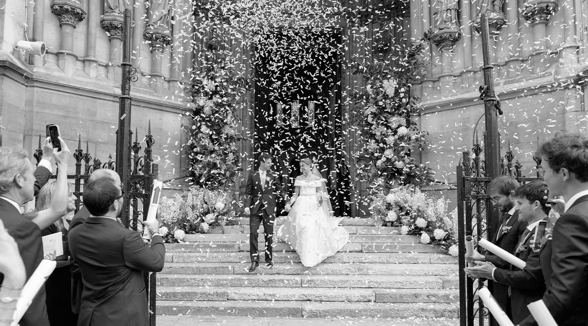 A bride and groom exit a church as guests shower them with confetti. The couple stands on the steps, surrounded by flowers and well-dressed people taking photos. The scene is joyful and festive.