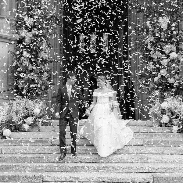 A bride and groom walk down the steps of a building, holding hands and smiling, surrounded by falling confetti and large floral arrangements on either side of the entrance. The scene is in black and white.