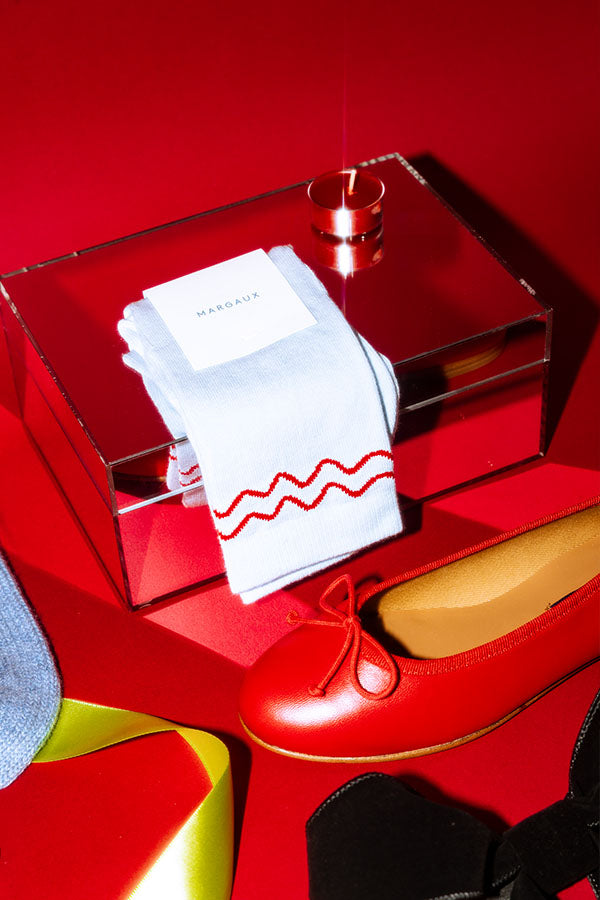 A pair of white socks with red wavy stripes and a Margaux label rest on a mirrored box, next to a red ballet flat shoe with a bow, against a bright red background.