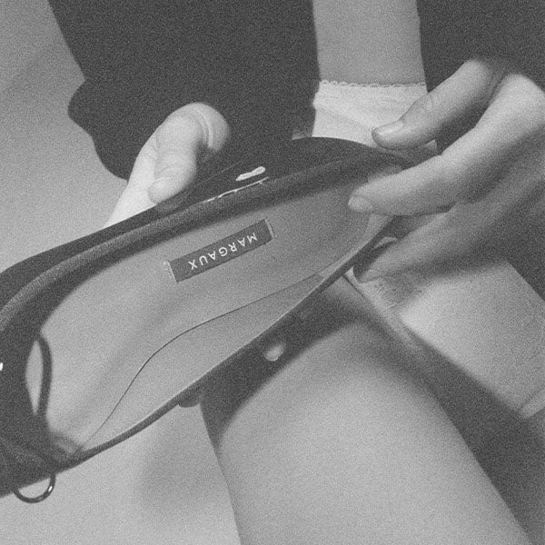 A person holds a shoe labeled “MARA DUX,” with one hand inside the shoe and the other hand on the heel. The image is in black and white, with a grainy texture.
