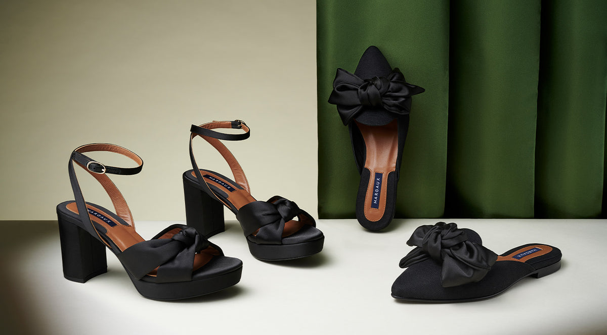 Three pairs of black women’s shoes with large bows: two platform heels with ankle straps and one pair of flat pointed-toe slip-ons, all displayed against a neutral and green background.