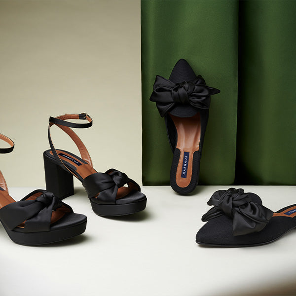 Three pairs of black women’s shoes with large satin bows are displayed: platform heels with ankle straps and block heels, and flat slip-on mules, all set against a green and beige backdrop.