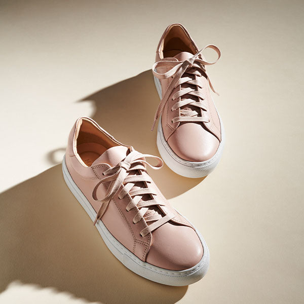 A pair of light pink sneakers with white soles and matching pink laces is placed on a beige surface, with soft shadows cast to one side.