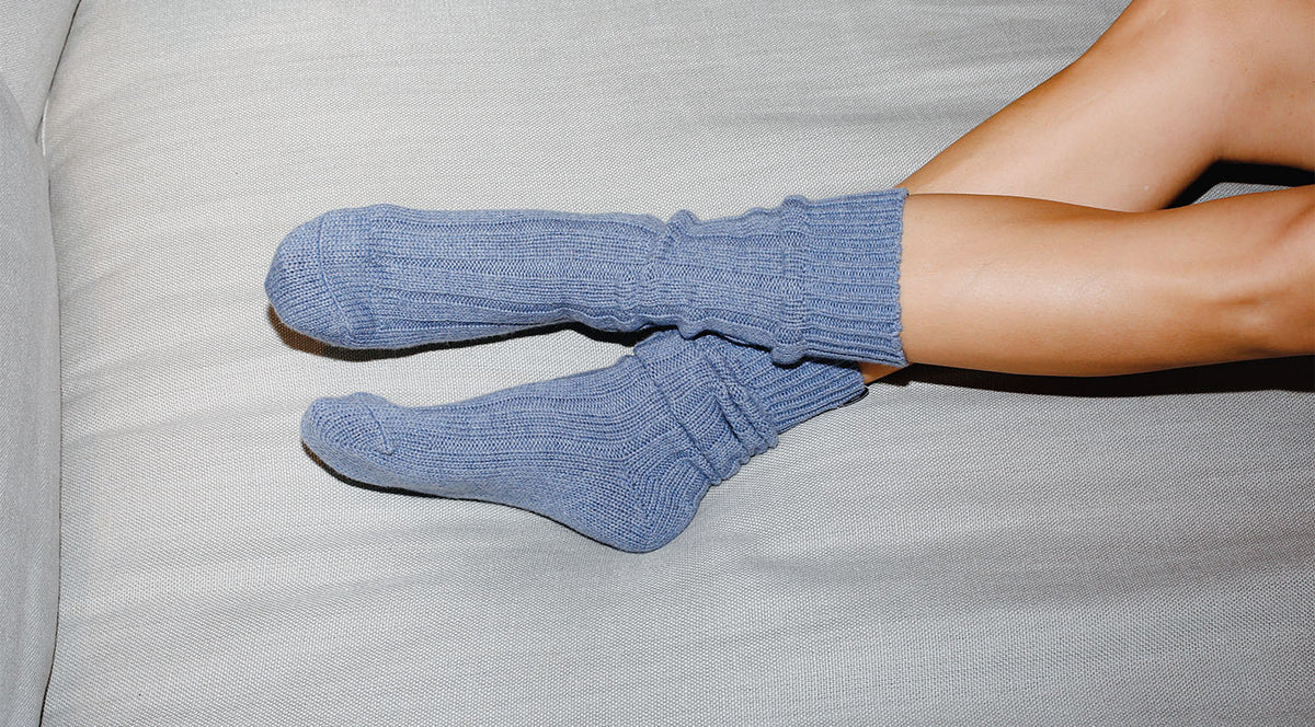 A person with bare legs is lying on a light gray couch, wearing thick, light blue knitted socks.