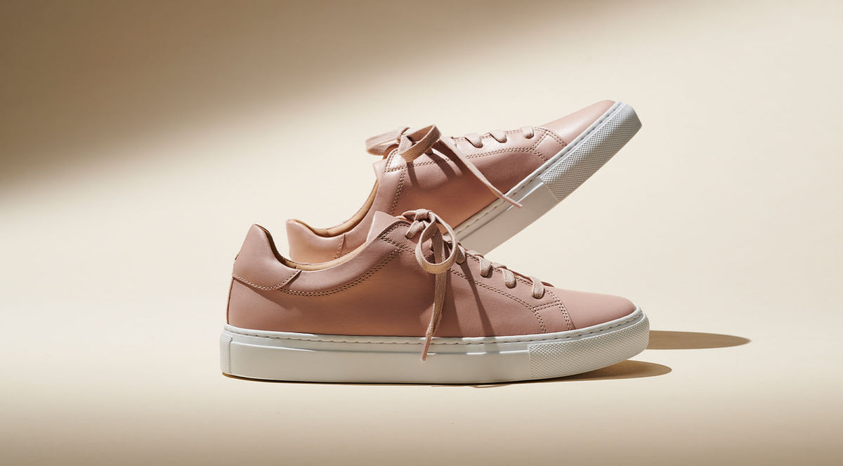 A pair of stylish light pink sneakers with white soles are arranged so that one shoe stands upright while the other is balanced on top, set against a beige background.