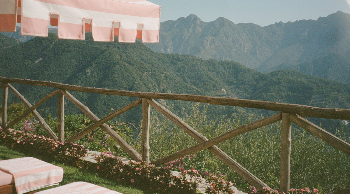 A wooden fence overlooks lush green mountains under a clear sky. Pink and white striped umbrellas and lounge chairs are in the foreground, with flowers lining the patio edge.