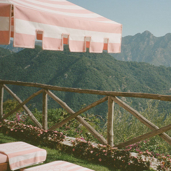 A pink-and-white striped umbrella shades lounge chairs beside a wooden fence, overlooking lush green mountains under a clear blue sky. Flowers line the edge of the patio area.