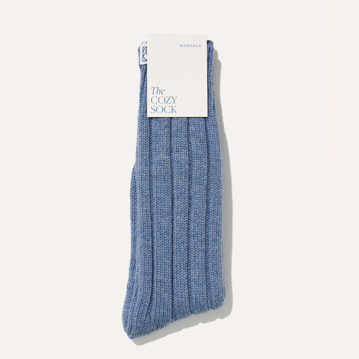 The Cozy Sock - Blue Cashmere by Margaux: a single blue ribbed sock in a soft wool-cashmere blend, featuring a white label with "The COZY SOCK" and "MARGAUX," set against a plain light background—ideal for luxurious lounging.