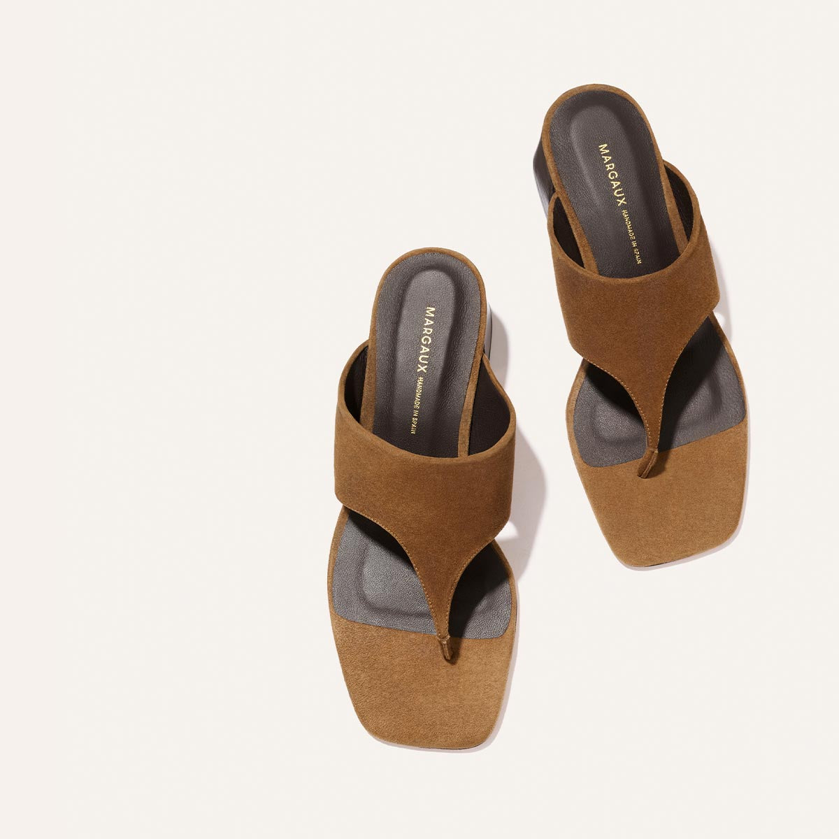 The Deia 50 - Nutmeg Suede by Margaux: brown suede thong sandals with a wide strap, square toe, black footbed, and "Margaux" in gold on the insole, shown against a light background.