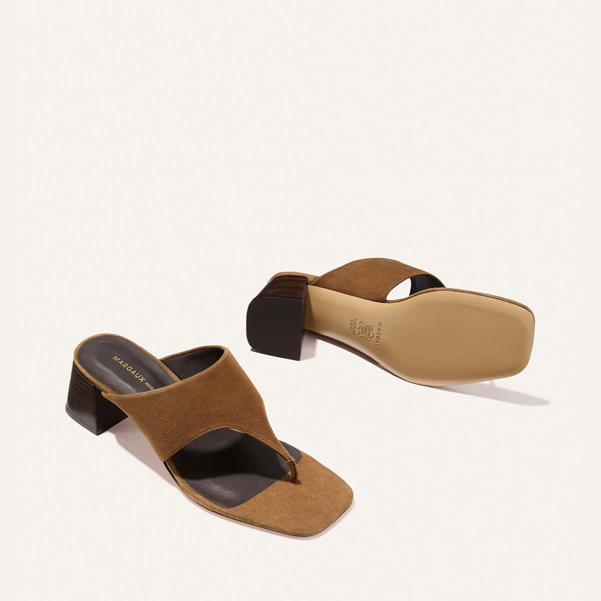 The Deia 50 - Nutmeg Suede by Margaux features brown suede heeled sandals with thick crisscross straps, a square toe, and block heels. One sandal stands upright; the other lies on its side, displaying the sole.