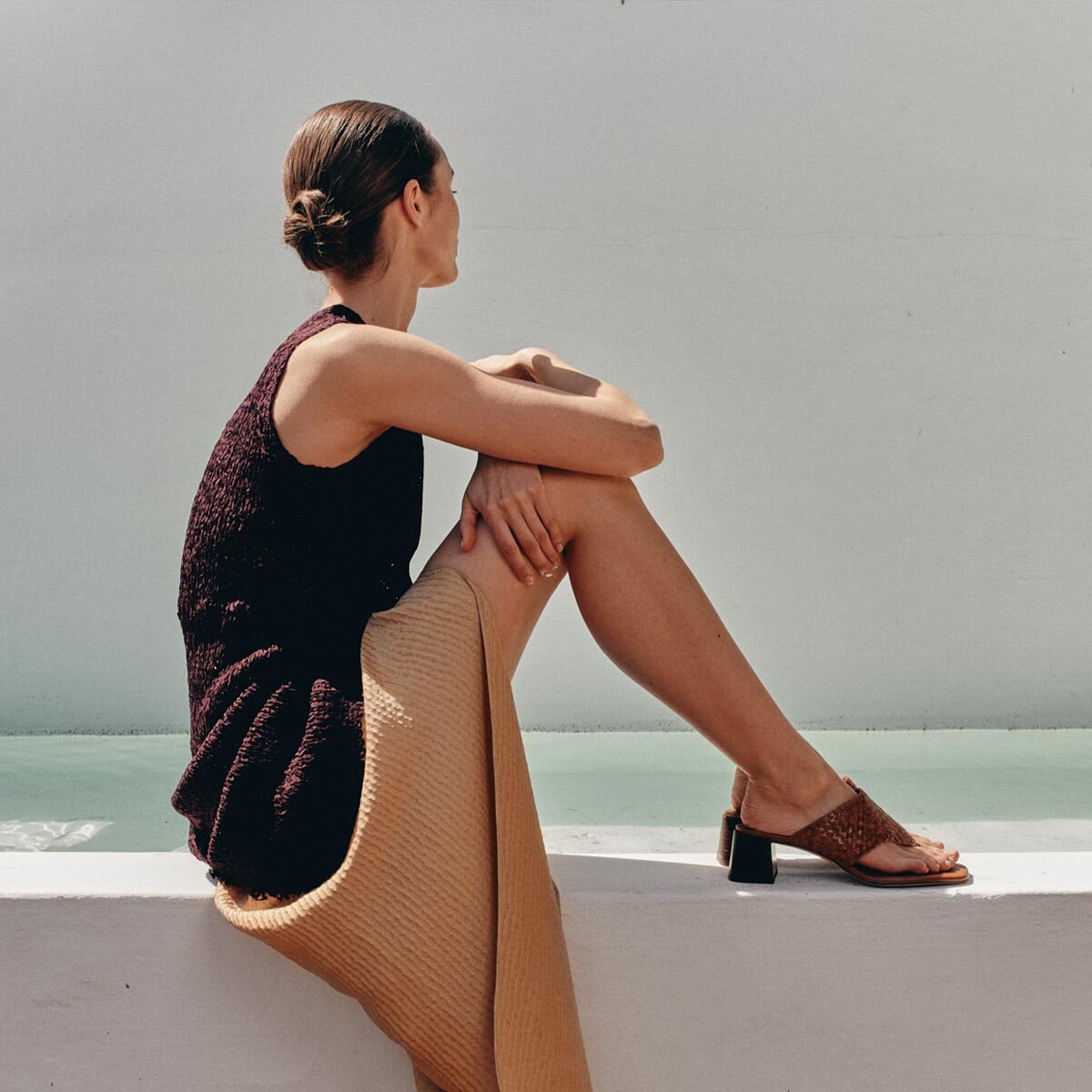 A woman with a low bun sits sideways on a white ledge, wearing a sleeveless dark top, tan skirt, and Margaux’s The Deia 50 - Saddle Woven Leather sandals, gazing thoughtfully into the distance against a pale background.