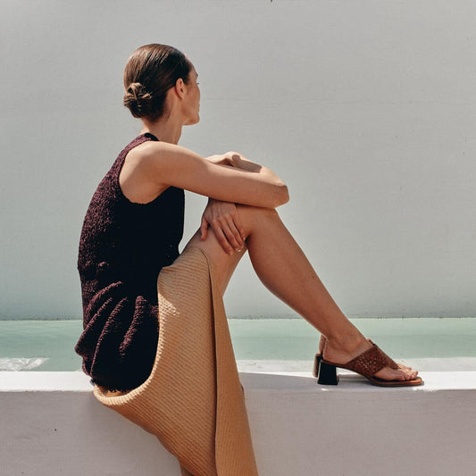 A woman with a low bun sits sideways on a white ledge, wearing a sleeveless dark top, tan skirt, and Margaux’s The Deia 50 - Saddle Woven Leather sandals, gazing thoughtfully into the distance against a pale background.