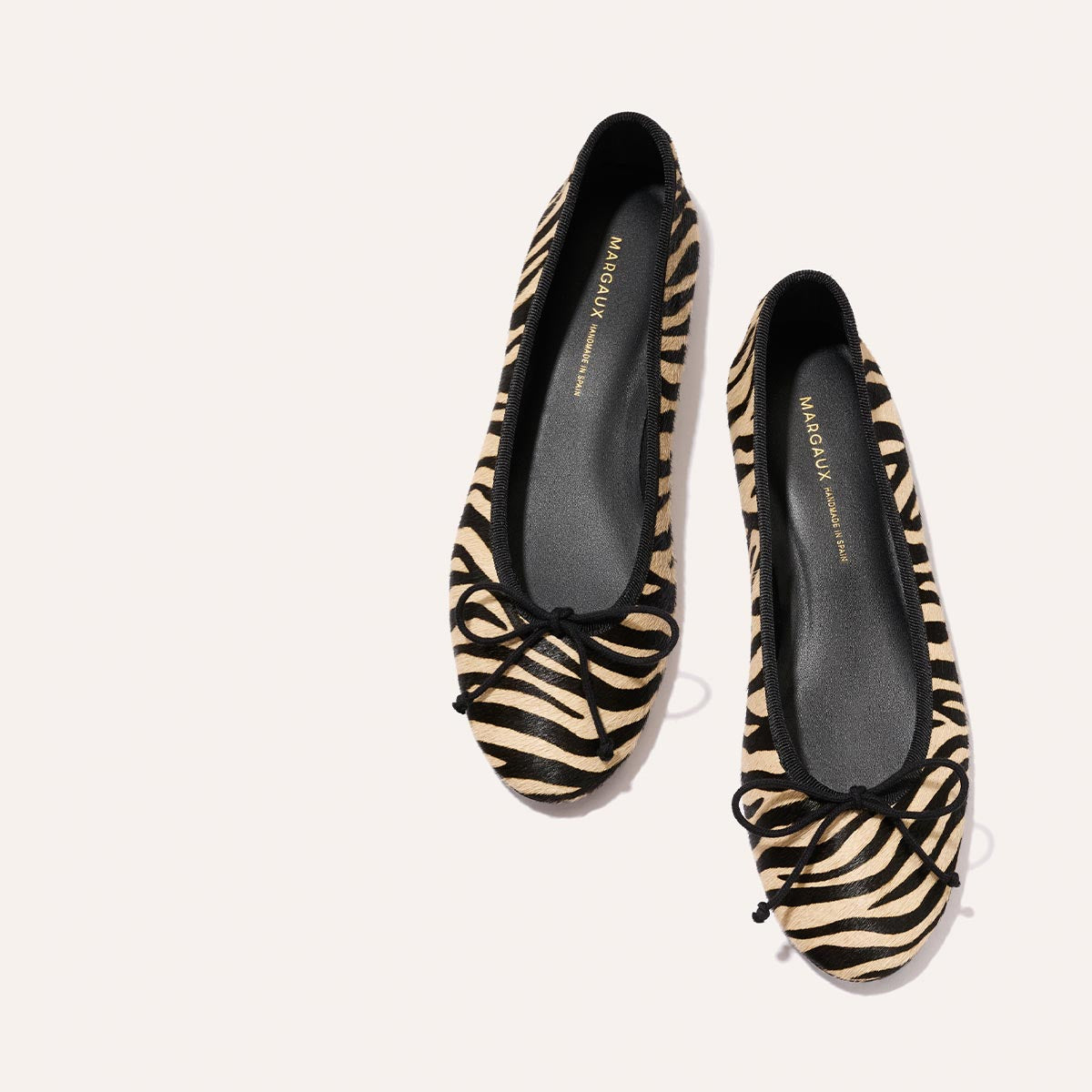 The Demi - Tan Zebra Haircalf ballet flats by Margaux, featuring comfortable zebra-print and black bow accents on the toes, are displayed against a light background.