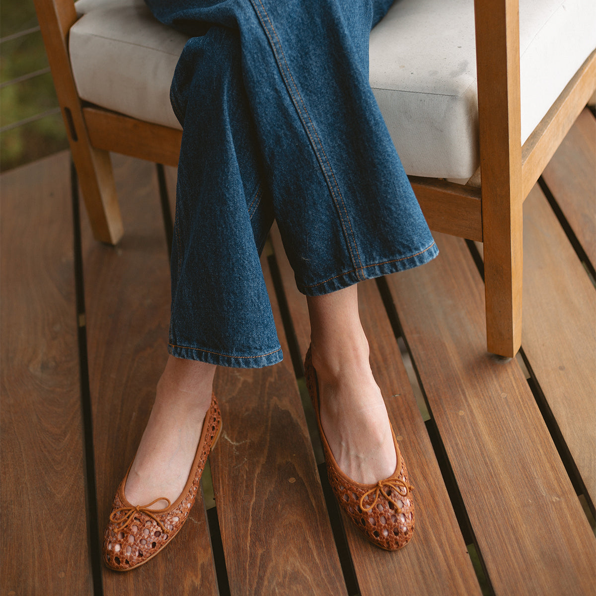 A person wearing blue jeans and The Demi - Saddle Woven Leather flats by margaux-development sits on a wooden chair with white cushioning, resting their feet on a wooden deck.