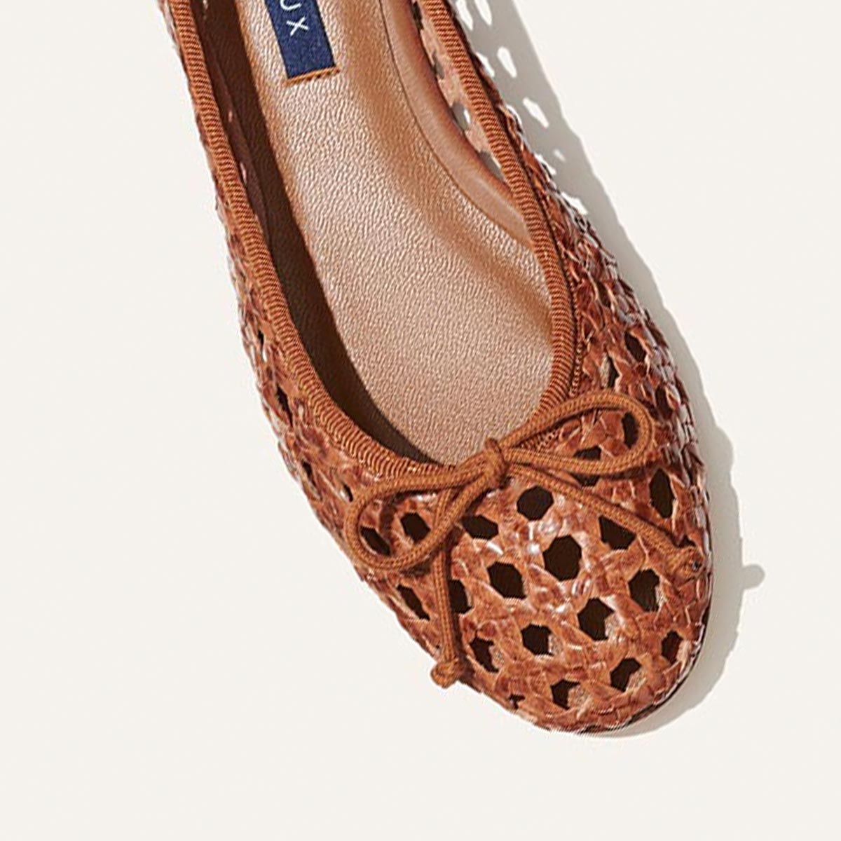 A close-up of The Demi - Saddle Woven Leather ballet flat by margaux-development, handmade in Spain with brown woven leather, a bow on the toe, and a beige insole, set against a light background.