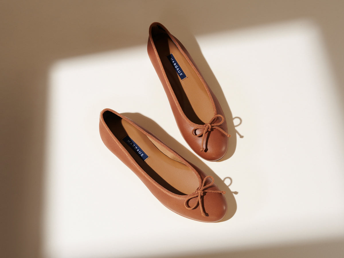 A pair of brown ballet flats with bows on the toes, placed on a light surface with soft shadows.