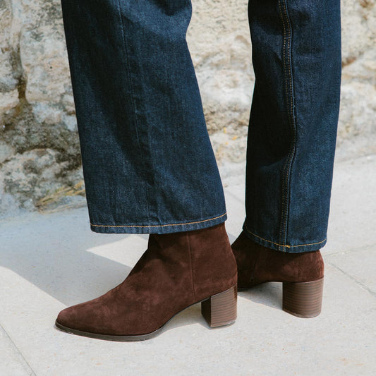 Someone wearing dark blue jeans and Margaux's The Downtown Boot First Edition in Chocolate Suede stands on light pavement near a stone wall.