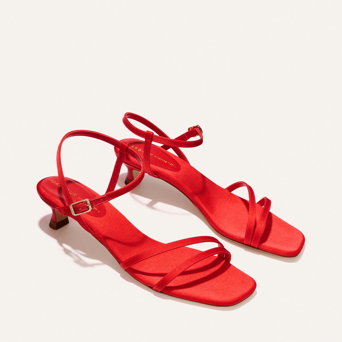 The Dylan Sandal by Margaux in Poppy Satin features a bright red hue, open toes, strappy design, low heels, and ankle straps, elegantly displayed on a plain white background.