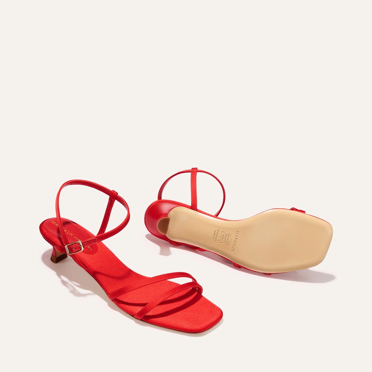 The Dylan Sandal in Poppy Satin by Margaux features bright red, strappy high heels with square toes and ankle straps—one upright showing the top, the other on its side to display the sole and heel.